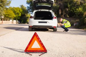 Triangle de présignalisation au premier plan suivi d'un conducteur qui répare sa voiture en second plan